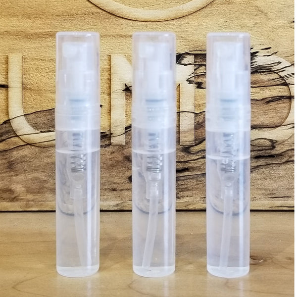 Three clear plastic spray bottles of Perfumology Sample Subscription Pack are arranged in a row in front of a wooden background etched with a bird design and partial text.
