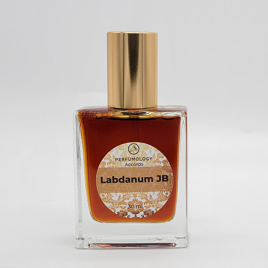 A dark amber liquid is housed in a rectangular glass bottle with a gold cap, labeled "Perfumology Accords: Labdanum JB by John Biebel, 30 mL.