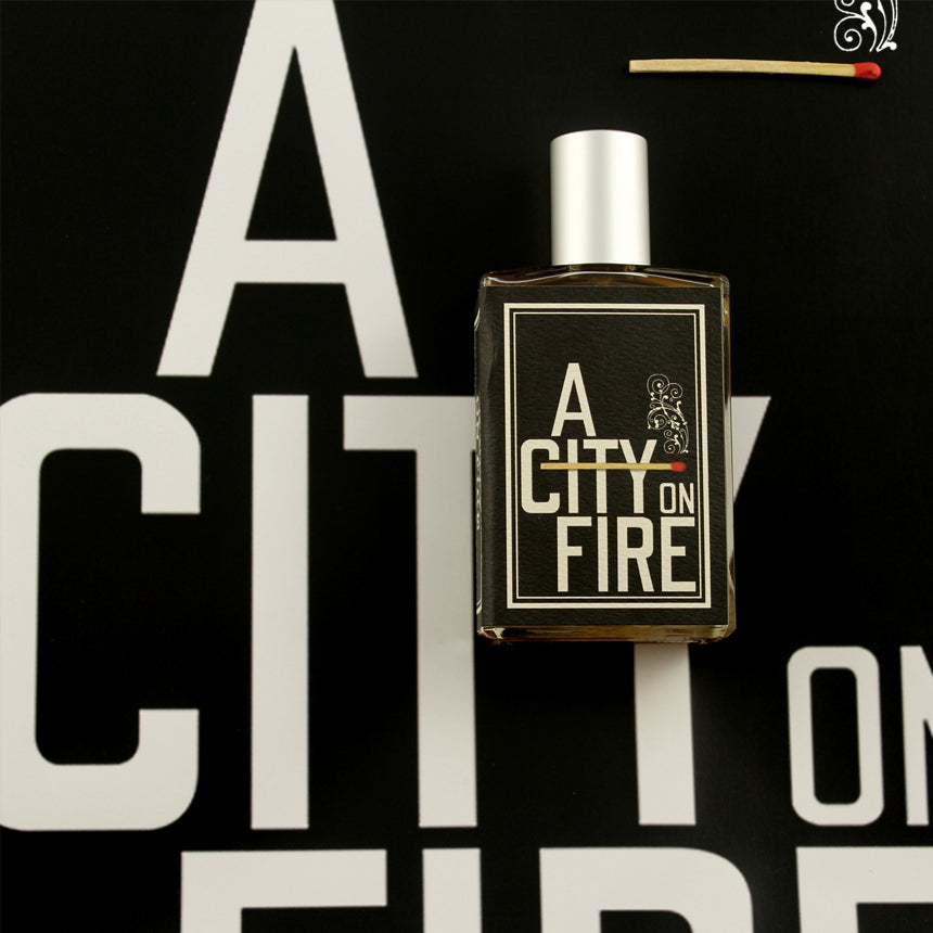 A bottle of "A City On Fire" from Imaginary Authors is placed on a matching background. A lit matchstick nearby hints at its refined smoke accord, capturing the essence perfectly.
