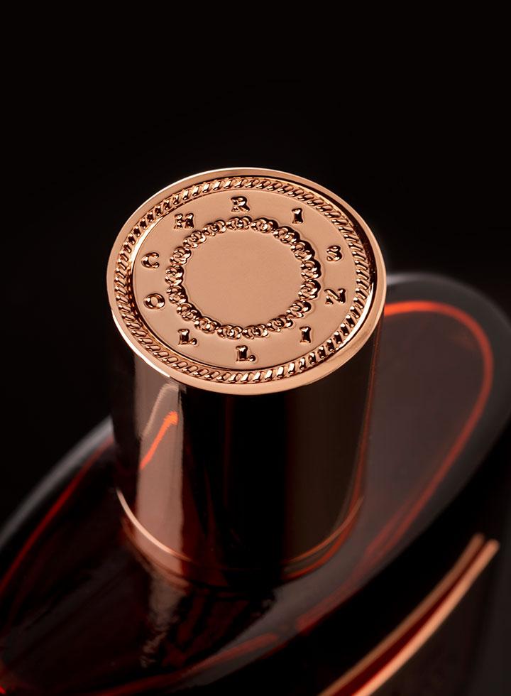 Close-up of a perfume bottle cap with embossed text around its circular edge. The cap has a metallic finish, and the bottle surface shows a sleek, dark design with subtle hints of red reflections—encapsulating the essence of cedarwood in this exquisite Chris Collins African Rooibos Eau de Parfum.