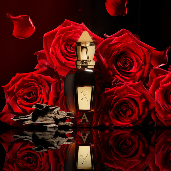 A perfume bottle with a gold cap is placed in front of red roses and wood logs, accompanied by floating rose petals, evoking the essence of Xerjoff's Alexandria Orientale oud perfume.