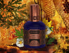 A dark blue and bronze bottle of Ambre Vie by House of Matriarch, an exquisite amber perfume known for its ultra-natural amber fragrance, is surrounded by flowers, greenery, and honeycomb in the background.