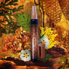 A cylindrical perfume bottle labeled "Ambre Vie" by House of Matriarch is surrounded by ultra-natural amber resin, flowers, greenery, honeycombs, and a sunset backdrop. The bottle cap is off and lies beside it.