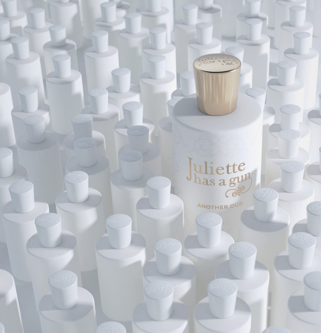 A single white bottle of "Another Oud" by Juliette Has A Gun with a gold cap stands out among numerous white bottles with white caps, each sharing the same design and shape, yet distinguished by its luxurious touch of oud wood.