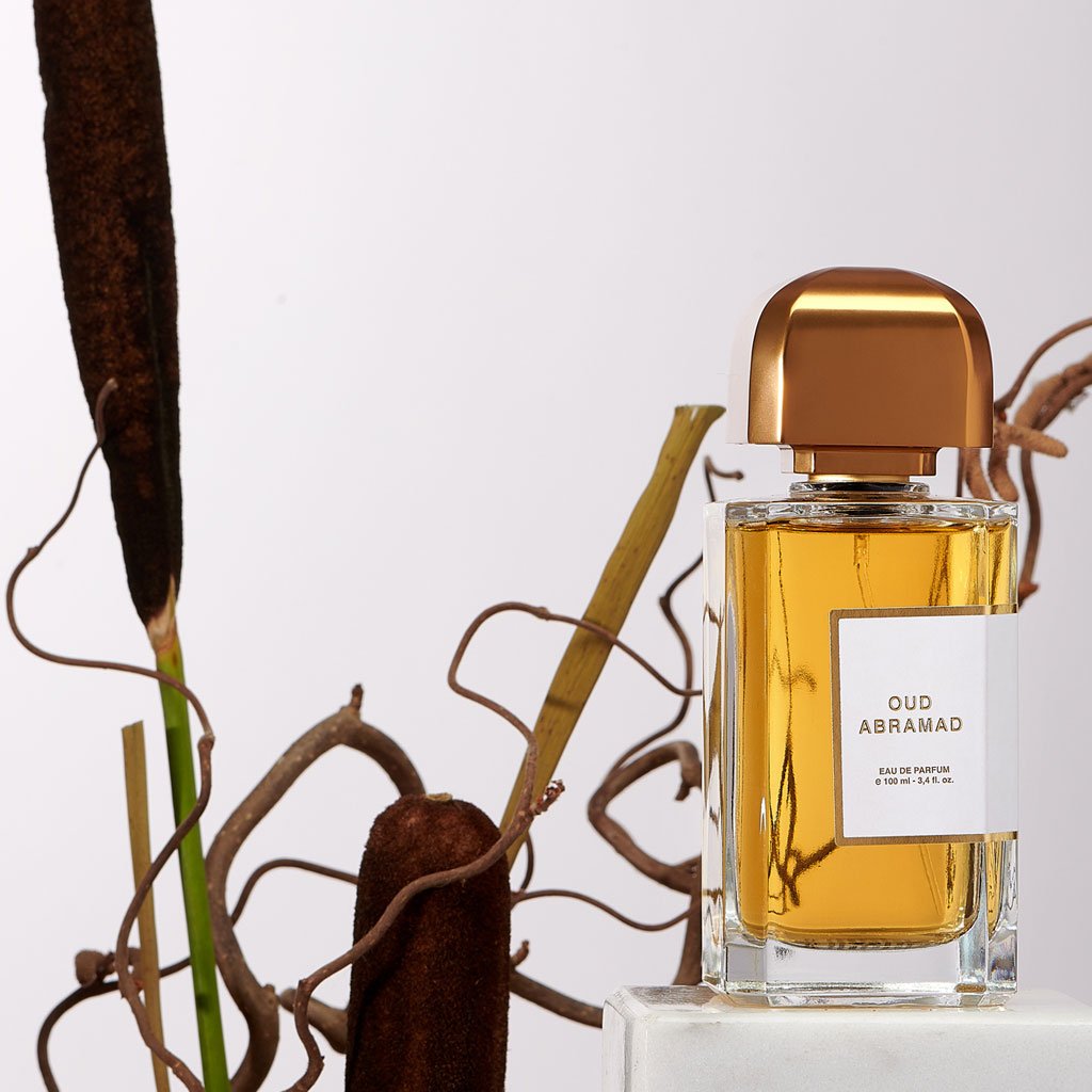 A BDK Parfums bottle labeled "Oud Abramad," adorned with a wooden cap, stands beside dried plant stems on a light background, evoking the mystique of the desert and the rich aroma of agarwood intertwined with Turkish rose.
