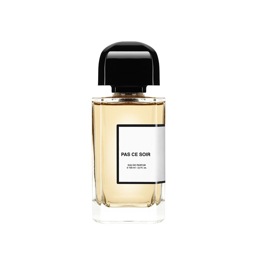 A clear glass bottle of "Pas Ce Soir" eau de parfum by BDK Parfums with a black cap, containing a light amber liquid, evocative of an evening stroll past Place de la Concorde, where the allure of red velour lipstick whispers secrets of Parisian cabaret nights.