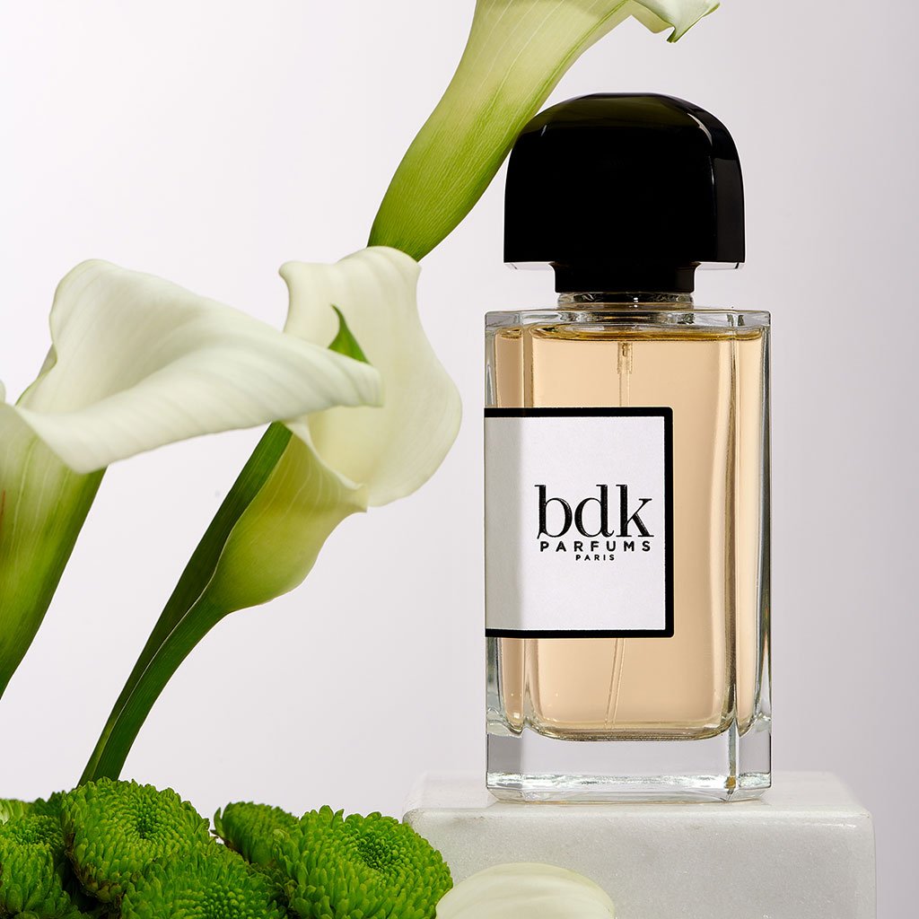 A bottle of BDK Parfums' Pas Ce Soir fragrance stands alongside white flowers and green decorative elements on a white background, evoking the allure of cabaret with the elegance of a Place de la Concorde evening.