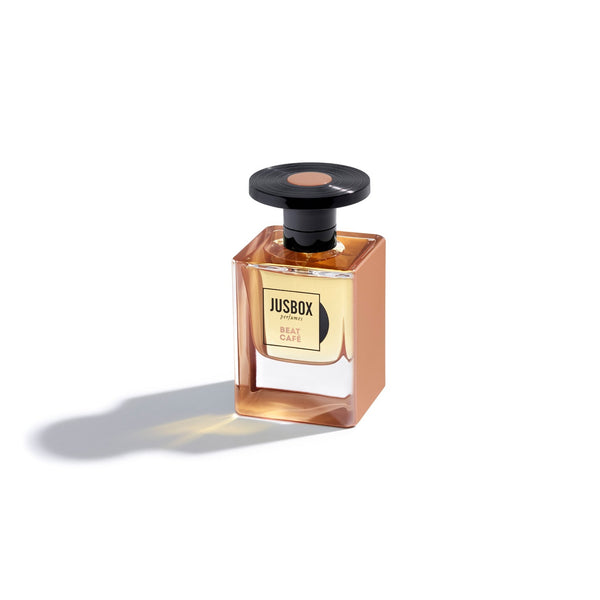 A small square glass bottle of Jusbox Beat Café perfume with a black round cap, casting a shadow on a white background, reminiscent of the Beat Generation era and echoing the vibes of 1962.