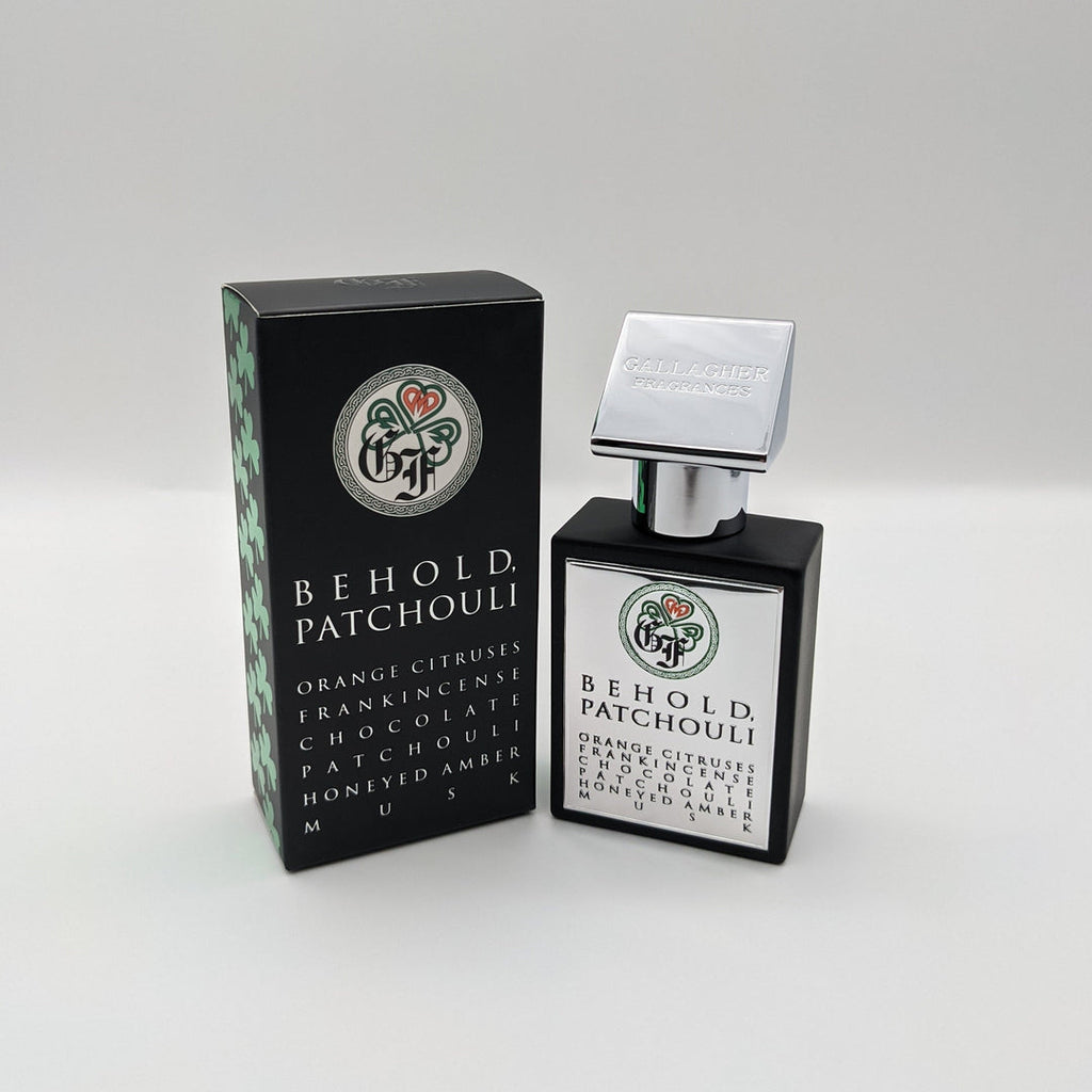 A black and silver perfume bottle labeled "Behold Patchouli" stands beside its black box packaging featuring green and white accents and a list of ingredients, a creation by renowned perfumer Daniel Gallagher for Gallagher Fragrances.