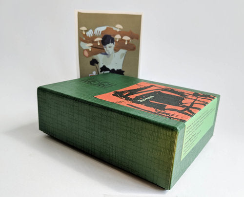 A green boxed set from January Scent Project, labeled "Burvuvu," with a visible cover image featuring an artistic illustration. In the background, there is a printed postcard showcasing another artwork of a person with hands raised, evoking notes of Eau de Parfum with hints of cedar and amber.