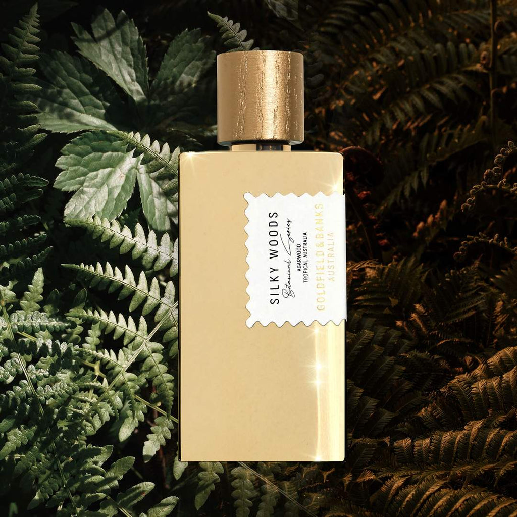 A bottle from Goldfield & Banks labeled "Silky Woods" sits among green and brown foliage, exuding the essence of a luxury fragrance with hints of agarwood.