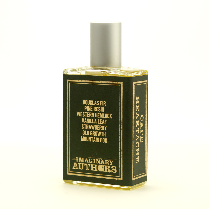 A rectangular glass perfume bottle labeled "Cape Heartache" by Imaginary Authors, featuring ingredient notes such as Douglas fir, pine resin, and vanilla leaf, evokes the mystique of the Pacific Northwest's old growth forests.