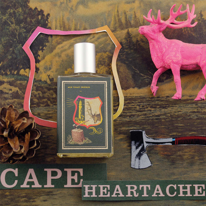 A bottle of "Cape Heartache" perfume by Imaginary Authors is surrounded by pinecones, an axe, a pink deer figurine, and a scenic Pacific Northwest forest background.