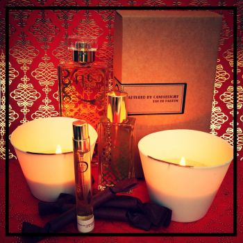 A display of perfume bottles, a box labeled "Captured by Candlelight" from 4160Tuesdays, lit candles in white holders, and wrapped chocolates against a red patterned background exuding rich fruity boozy notes.