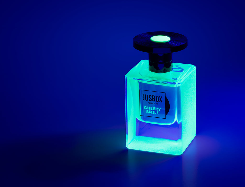 A square glass bottle of Jusbox's "Cheeky Smile" perfume is bathed in a blue glow, evoking the energetic essence of Acid House, set against a dark backdrop.