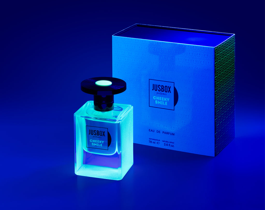 A Jusbox Cheeky Smile perfume bottle is elegantly positioned in front of its fluorescent packaging, with the scene enhanced by a blue lighting effect.