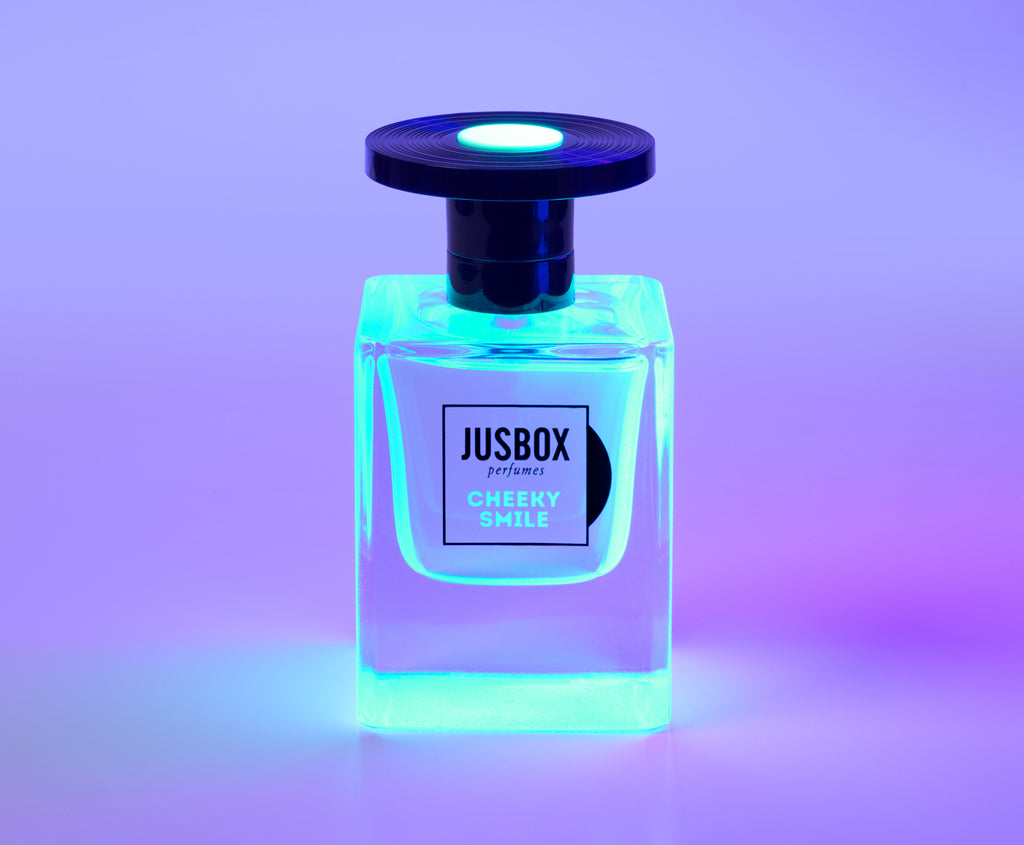 A bottle of Jusbox's "Cheeky Smile" fragrance, its fluorescent packaging illuminated with blue and purple lighting against a gradient background.