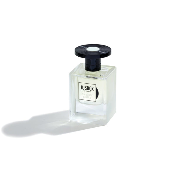 A transparent glass bottle of Jusbox's "Cheeky Smile" perfume, adorned with a sleek black cap, casts a gentle shadow on a pristine white background, radiating the vibrant energy of Acid House through its unique molecular fragrance.