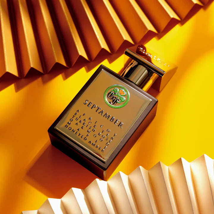 A rectangular perfume bottle labeled "Septamber" by Gallagher Fragrances is artfully placed on a bright yellow background, accented with corrugated paper. The label highlights captivating notes of black rose, vanilla, oud, and honeyed amber—ideal for Fall and Winter seasons.