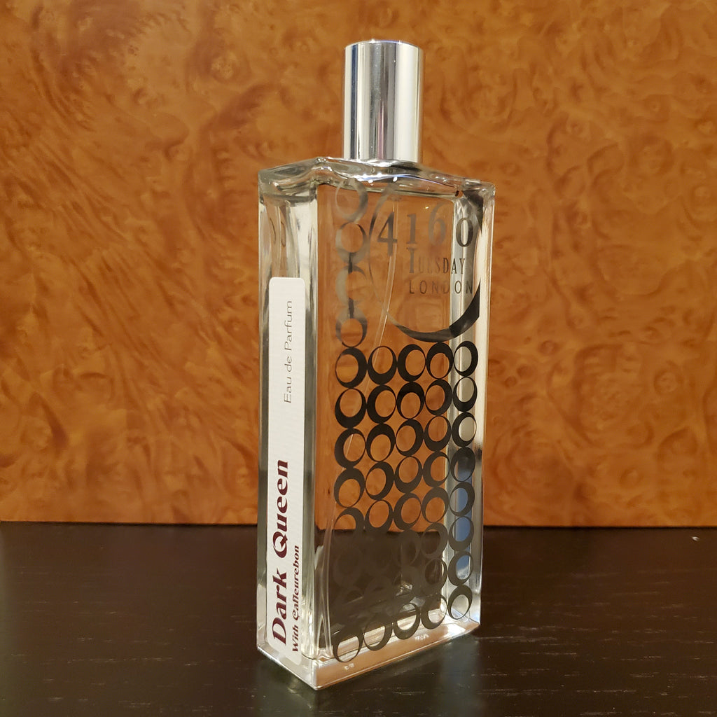 A clear glass perfume bottle labeled "4160Tuesdays" with a pattern of black circles and a separate label reading "Dark Queen Eau de Parfum," resting on a dark surface, exudes an aura befitting both Dark Queen and White Queen.