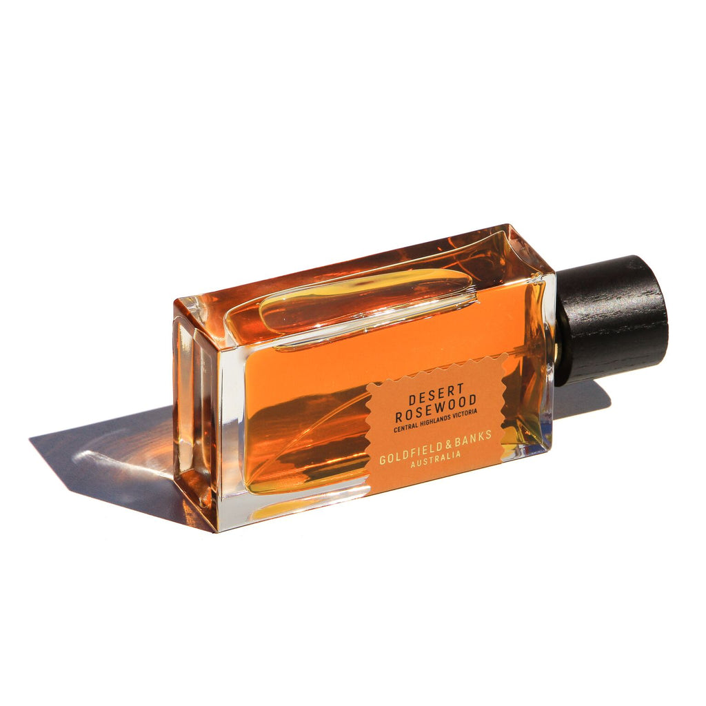 A rectangular glass bottle with a black cap contains an amber-colored liquid. The label reads, "Desert Rosewood" by Goldfield & Banks, Australia. This luxurious bottle holds a leathery fragrance enriched with notes of wood and spice.