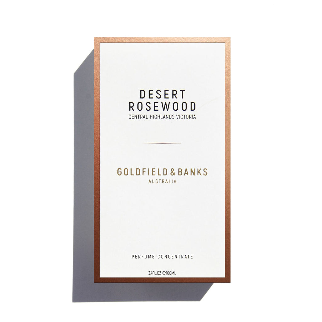 A rectangular white box with copper edges displays the text "Desert Rosewood" and "Goldfield & Banks" in black and gold. It contains a wood and spice perfume concentrate, offering a leathery fragrance, with 3.4 FL OZ (100mL) indicated at the bottom.