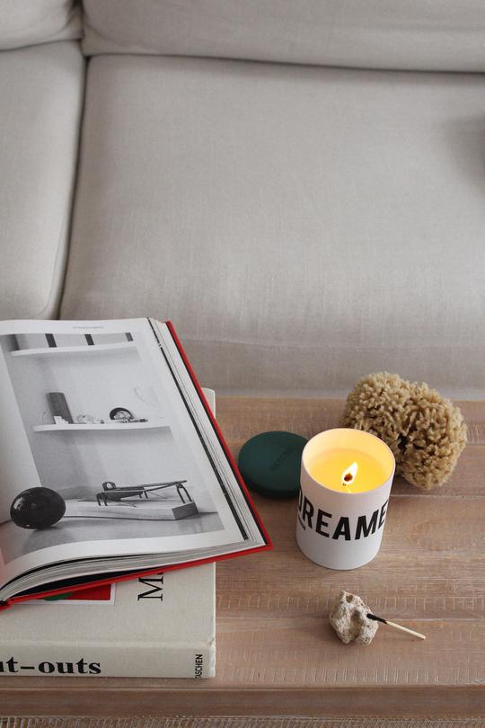 A wooden table with an open book, a lit Nomad Noe candle labeled "DREAMER in London," a piece of coral, and a shell. A beige sofa is in the background.