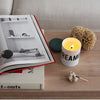 A wooden table with an open book, a lit Nomad Noe candle labeled "DREAMER in London," a piece of coral, and a shell. A beige sofa is in the background.