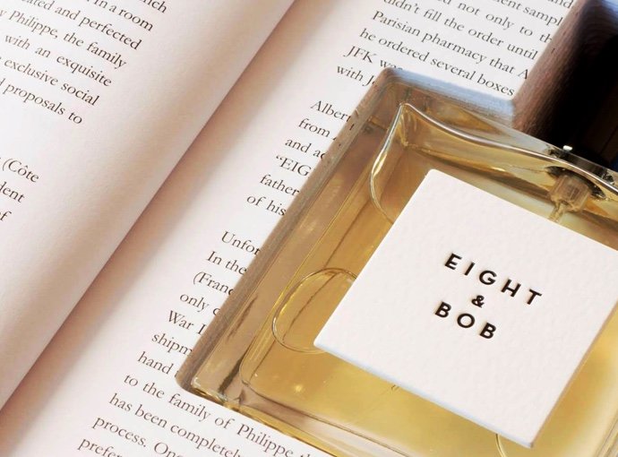 A bottle of Eight & Bob's Eight & Bob exclusive cologne rests on an open book, partially covering the text on the pages.