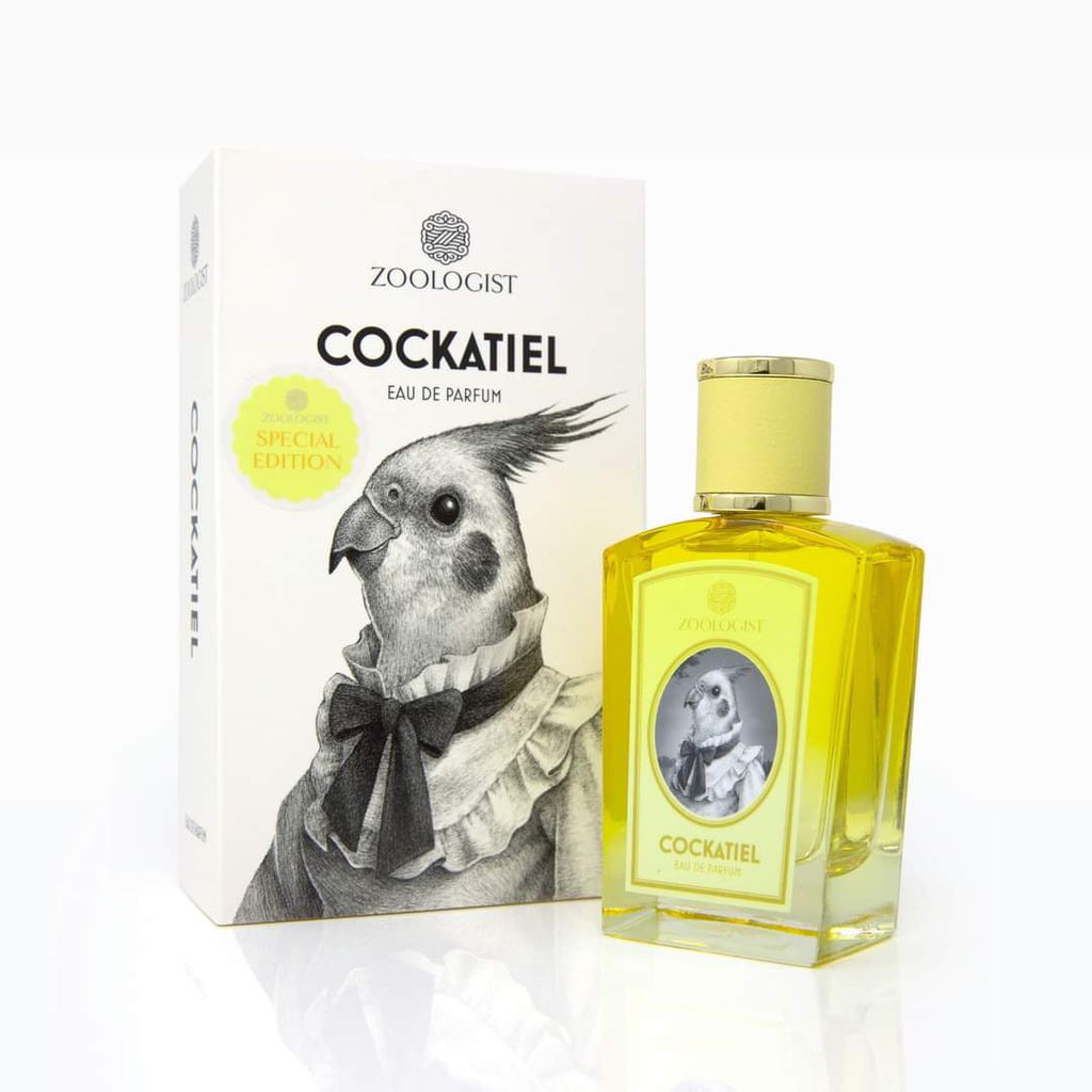 A bottle of Zoologist Cockatiel is placed next to its matching box, which features a drawing of a cockatiel wearing a tie. The yellow liquid inside the bottle evokes an Australian spring, with hints of yellow acacia adorning the label’s similar image.