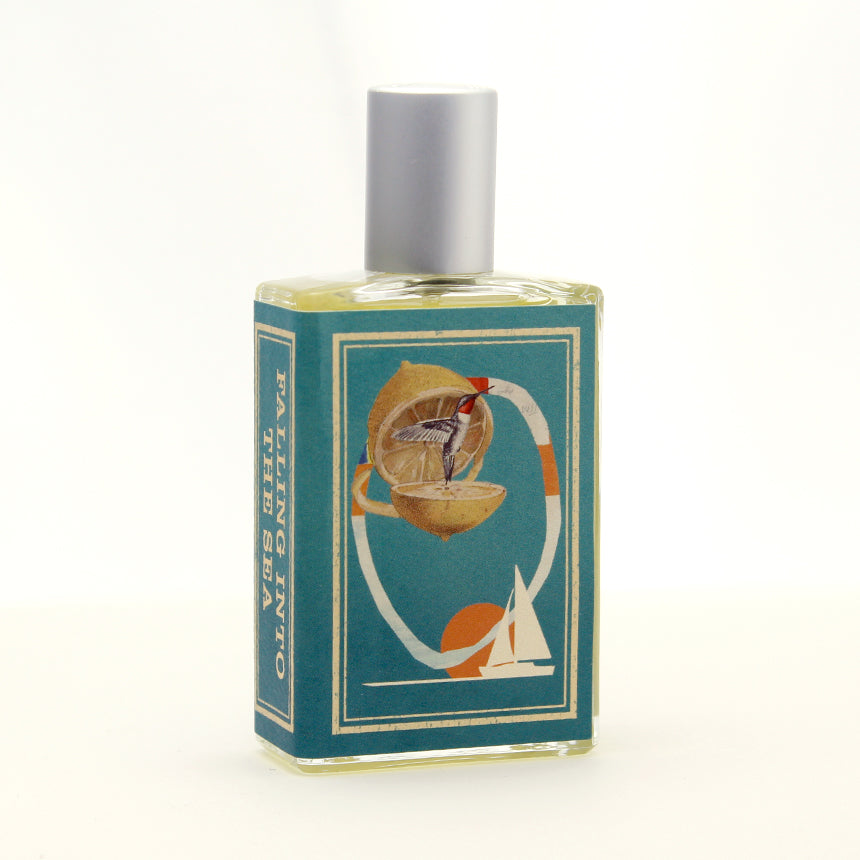 A rectangular perfume bottle with an illustration of a fruit and a ship on the label. The text on the label reads "FALLING INTO THE SEA." The bottle, designed by Josh Meyer, features a silver cap and contains a yellow liquid that captures the essence of a refreshing summer scent. This is *Falling Into the Sea* by *Imaginary Authors*.