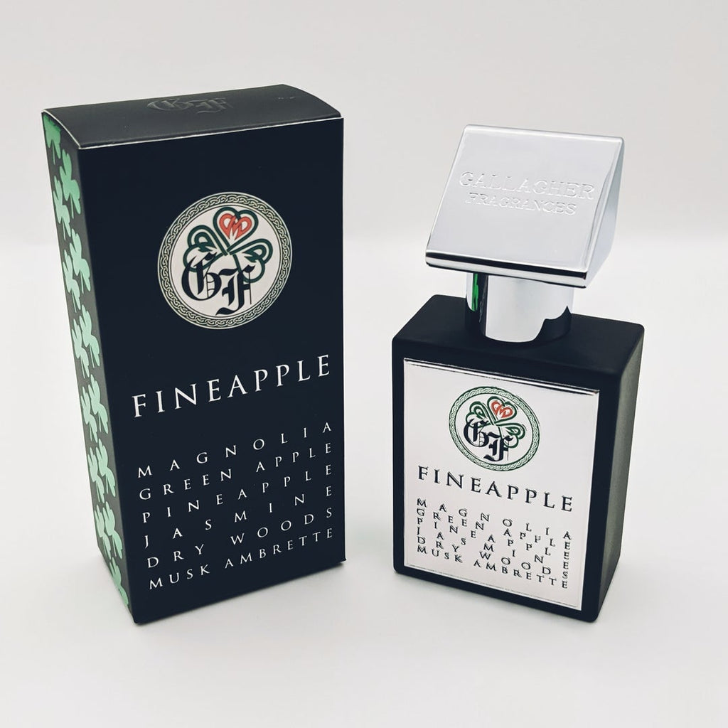 A black and white bottle of Fineapple perfume by Gallagher Fragrances, adorned with a decorative logo, stands beside a classy black and green box featuring the same elegant branding. The packaging highlights the enchanting notes of Magnolia, Green Apple, Pineapple, Jasmine, Dry Woods, and Musk Ambrette.