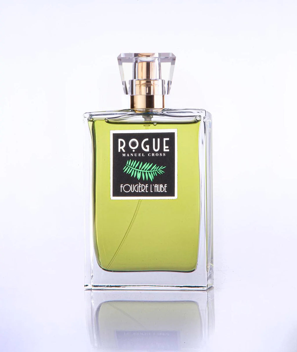 A clear glass bottle of Rogue Perfumery's "Fougere L'Aube" fragrance reveals a green citrus-infused, greenish-yellow liquid inside, topped with a gold-colored cap and a transparent cover.