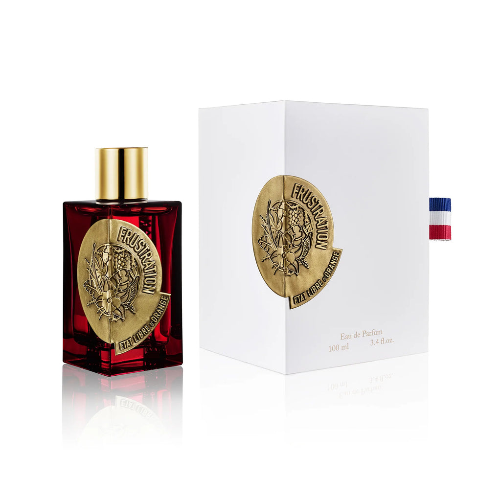 Frustration EDP by État Libre d'Orange, a red glass bottle of perfume with a gold cap and emblem, sits next to a white box featuring the same emblem. The box includes text and is adorned with a ribbon sporting red, white, and blue stripes. The fragrance inside harmoniously blends notes of vanilla and vetiver for an elegant finish.