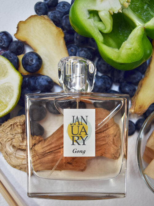 A clear glass bottle labeled "January Scent Project Gong" is surrounded by blueberries, a lemon slice, a green bell pepper, ginger, dried fruit, and wooden sticks. The enticing aroma suggests notes of Fruit and Green Musk, capturing the essence of nature's bounty in a refined perfume.