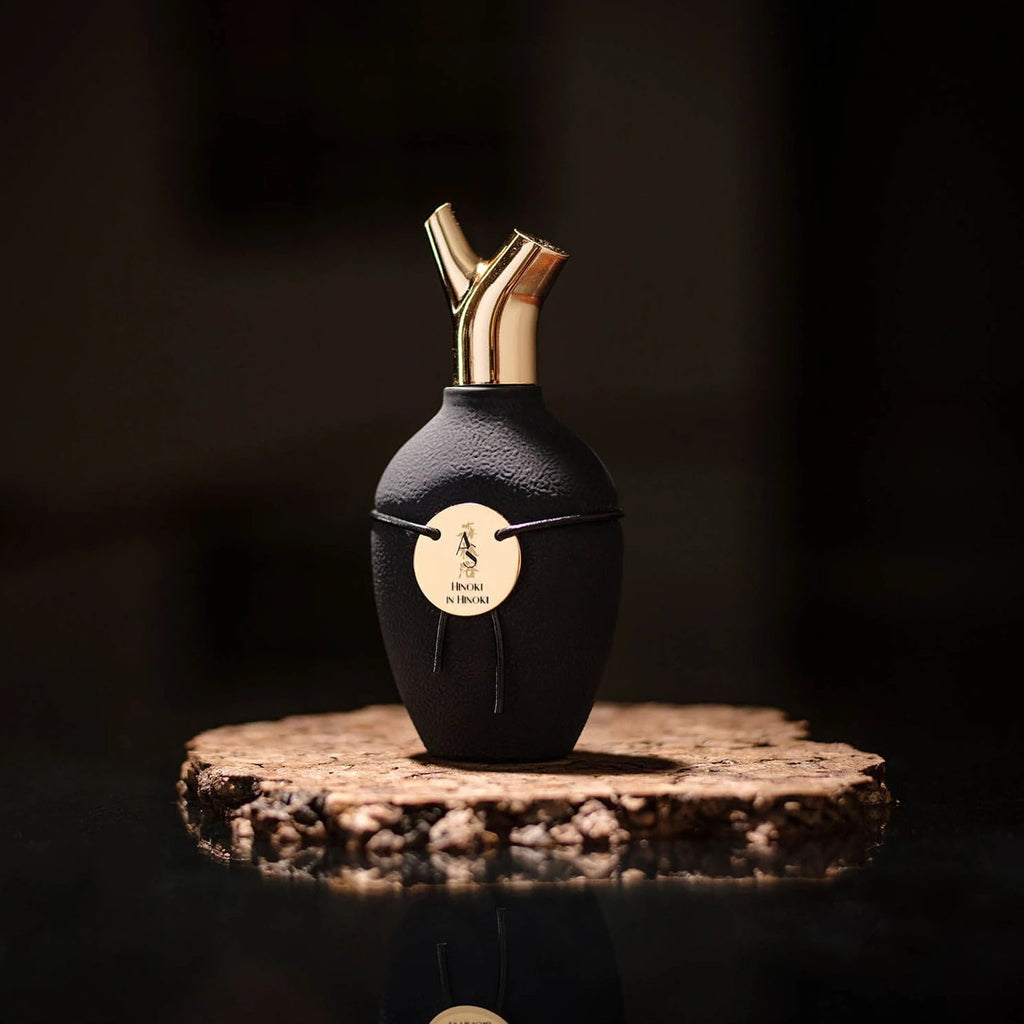 A black perfume bottle with gold dual spouts and a round label rests on a wooden slab against a dark background, exuding the essence of Hinoki in Hinoki by Scents of Wood.