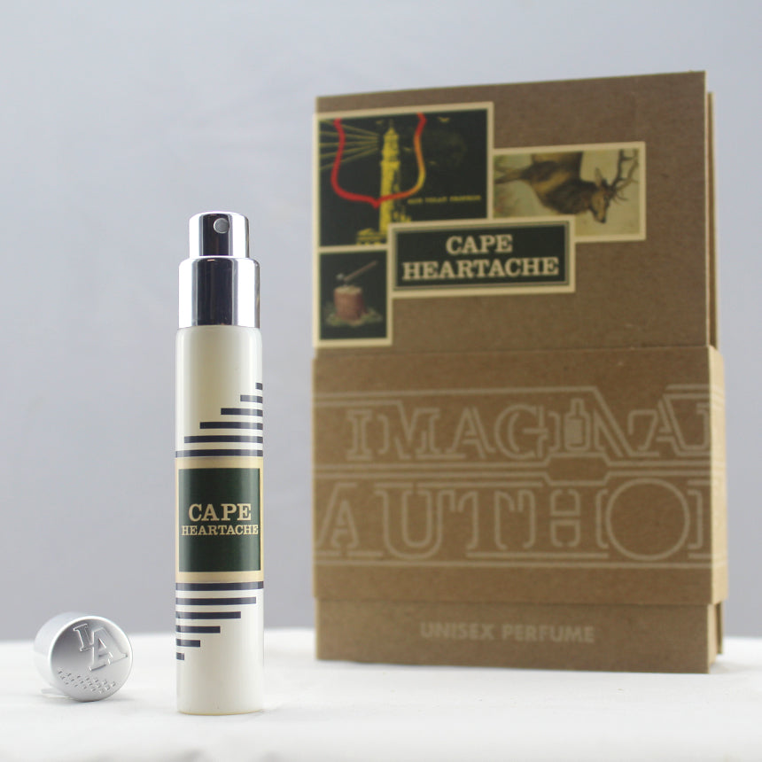 A bottle of Cape Heartache unisex perfume by Imaginary Authors, positioned in front of its packaging box. The Cape Heartache bottle is white with a black and gold label, evoking the essence of old growth forests in the Pacific Northwest. The cap is placed beside it.