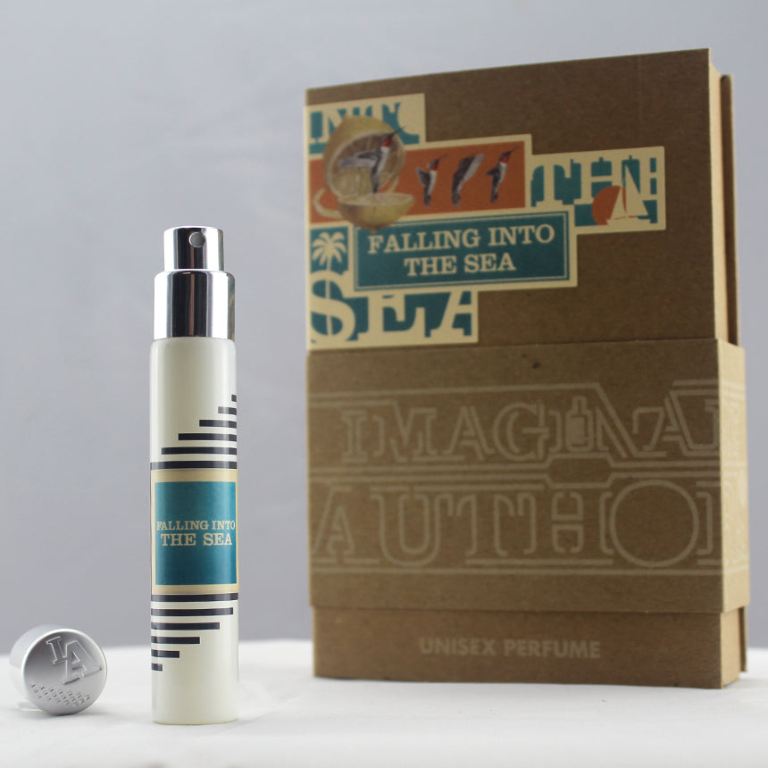 A small bottle of "Falling Into the Sea" unisex perfume, created by Imaginary Authors, is placed next to its decorated box. The bottle has a striped design that hints at its refreshing summer scent with notes of lemon. The box features colorful text and graphics, capturing the essence of sunshine and waves.