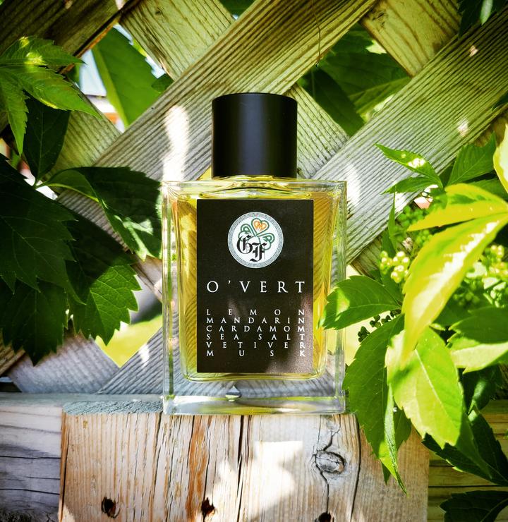 A bottle of O'Vert by Gallagher Fragrances rests on a wooden surface, set against a wooden lattice with green leaves and plants in the background. The label lists lemon, green mandarin, cardamom, sea salt, vetiver, and musk.