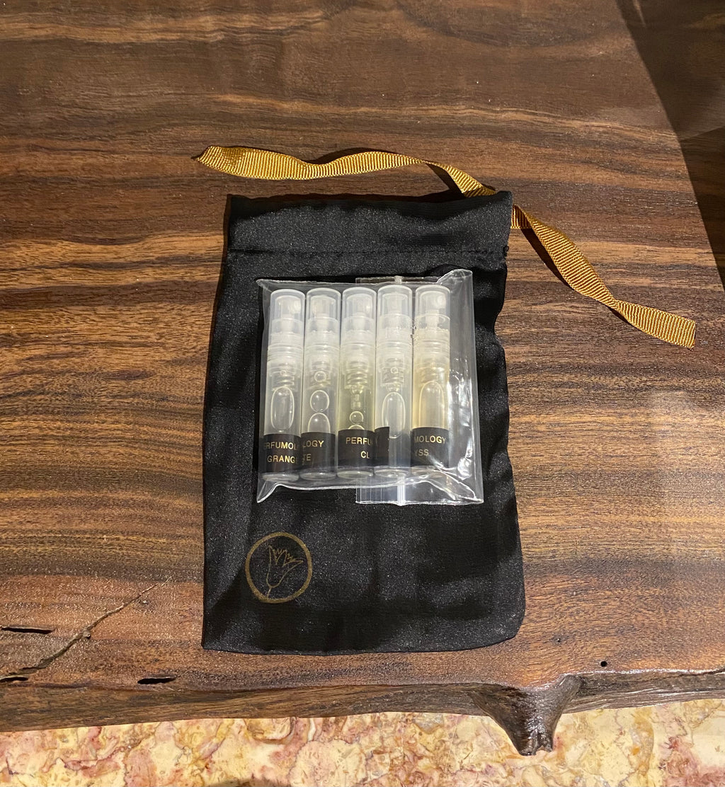 The Perfumology Discovery Set by Perfumology features a small black silk baggie with a golden ribbon, containing a clear plastic case that holds six small labeled perfume samples. Both the elegant silk bag and the bottles are showcased on a wooden surface.