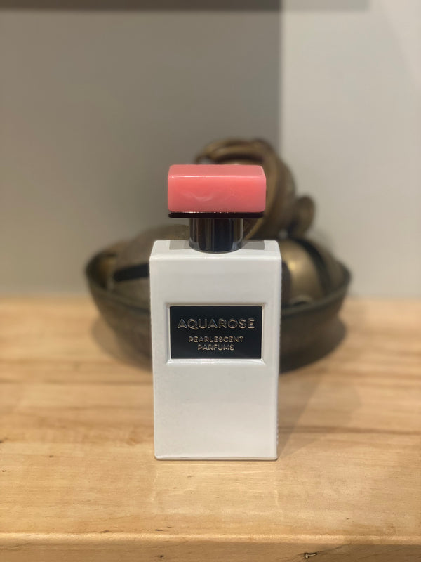 A bottle of Aquarose by Pearlescent Parfums with a white body and pink cap, placed on a wooden surface against a neutral background.