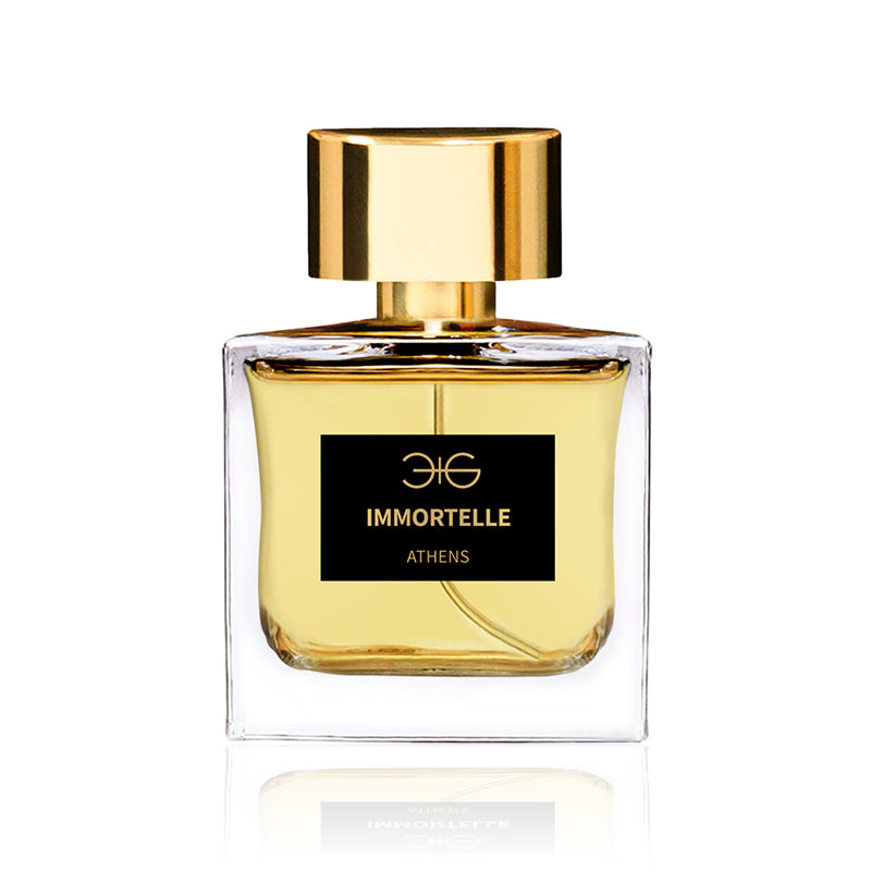 A rectangular glass bottle of Immortelle Manos Gerakinis perfume, with a gold cap and label showcasing the brand and fragrance name, reflects its Greek heritage. The bottle contains a fragrant golden-yellow liquid.