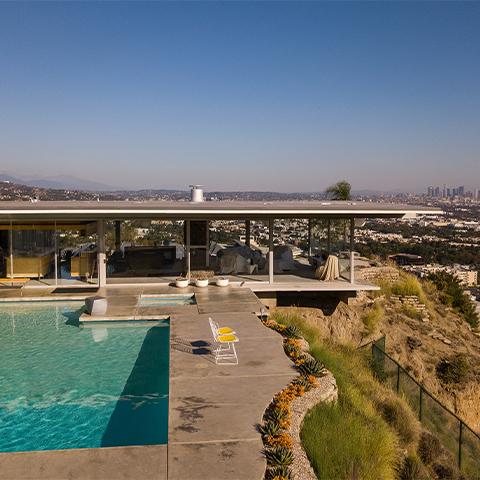 A modern house with large glass windows overlooks a cityscape. The property features an outdoor pool, patio area, and surrounding vegetation that sways gently in the marine breeze of Los Angeles by Gallivant from Gallivant Perfumes.