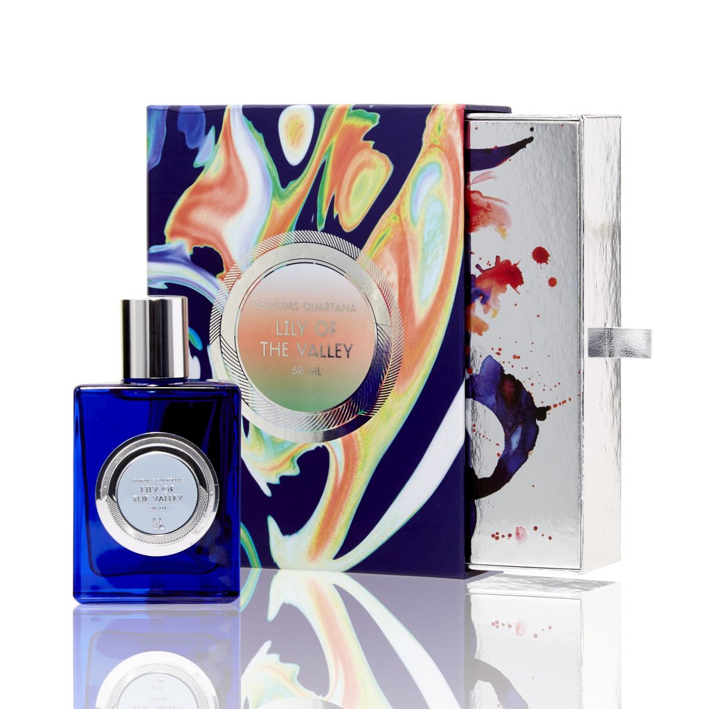 A blue perfume bottle labeled "Lily of the Valley" from Parfums Quartana stands in front of an abstractly designed, colorful box. The silver lid is partially open beside it, hinting at the delicate scents of Lily of the Valley and a subtle black leather accord within this Eau de Parfum.