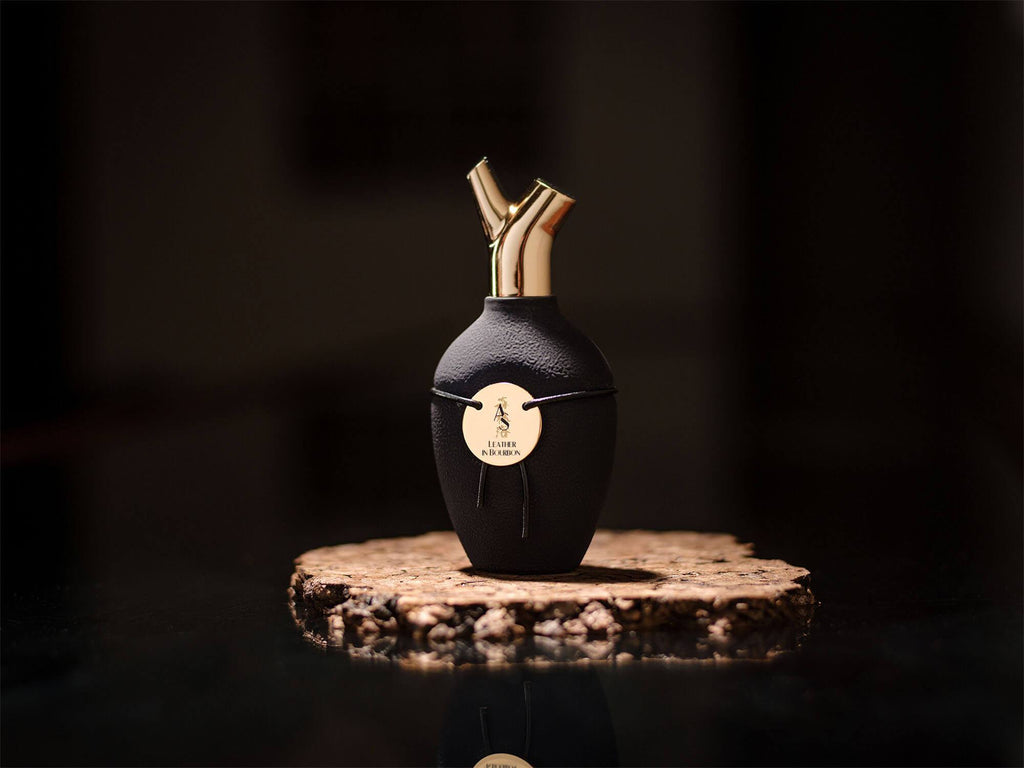 A bottle of Leather In Bourbon by Scents of Wood, characterized by gold-tipped dual spouts and a round gold label, exuding a luxurious fragrance, is displayed on a round wooden base against a dark background.
