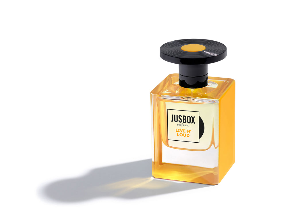 A bottle of "Live 'N' Loud" by Jusbox, featuring yellow liquid and a black cap, casts a shadow on a white background. Inspired by music, this fragrance blends notes to uplift mental well-being.