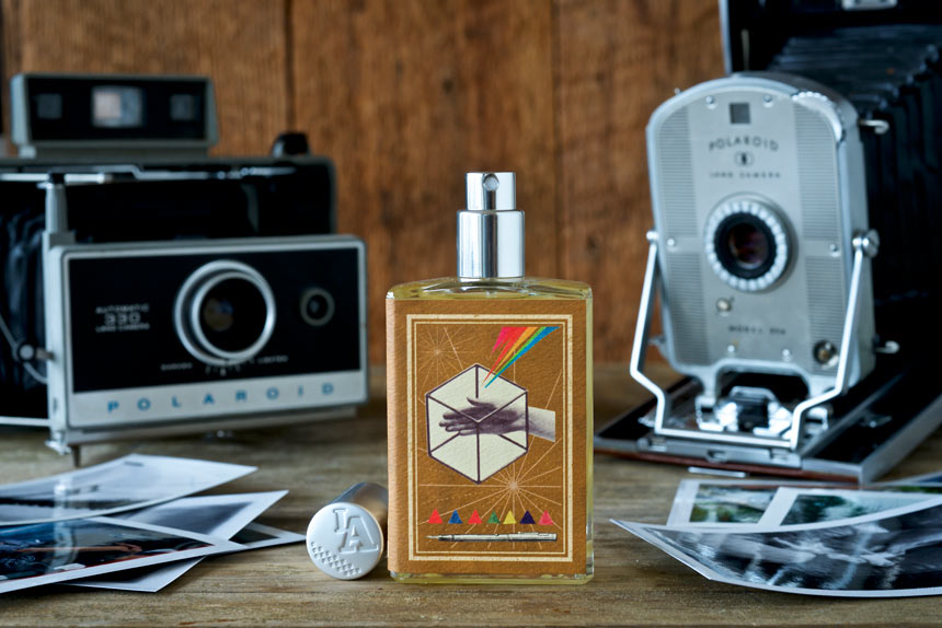 A bottle of Memoirs of a Trespasser cologne by Imaginary Authors, infused with the essence of Madagascar Vanilla, stands on a wooden surface flanked by vintage Polaroid cameras and scattered photos.