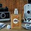 A bottle of Memoirs of a Trespasser cologne by Imaginary Authors, infused with the essence of Madagascar Vanilla, stands on a wooden surface flanked by vintage Polaroid cameras and scattered photos.
