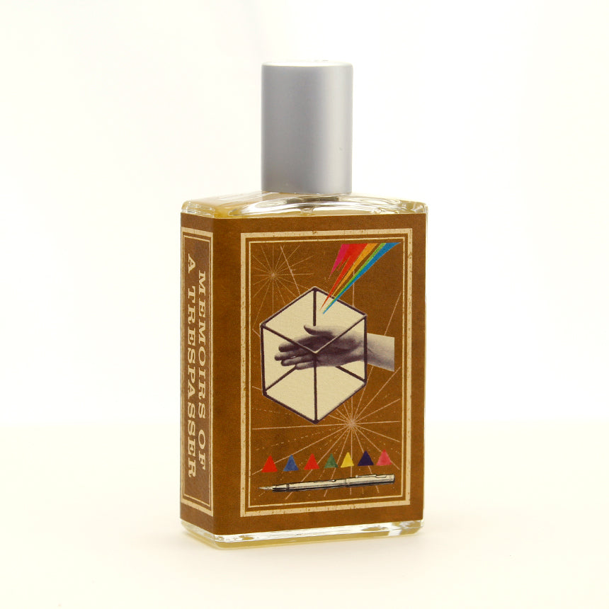 A rectangular bottle with a silver cap contains a brown liquid. The label, bearing the name "Memoirs of a Trespasser" by Imaginary Authors, features an abstract design of a hand within a geometric shape and colorful beams, evoking an adventurous scent with hints of Madagascar Vanilla.