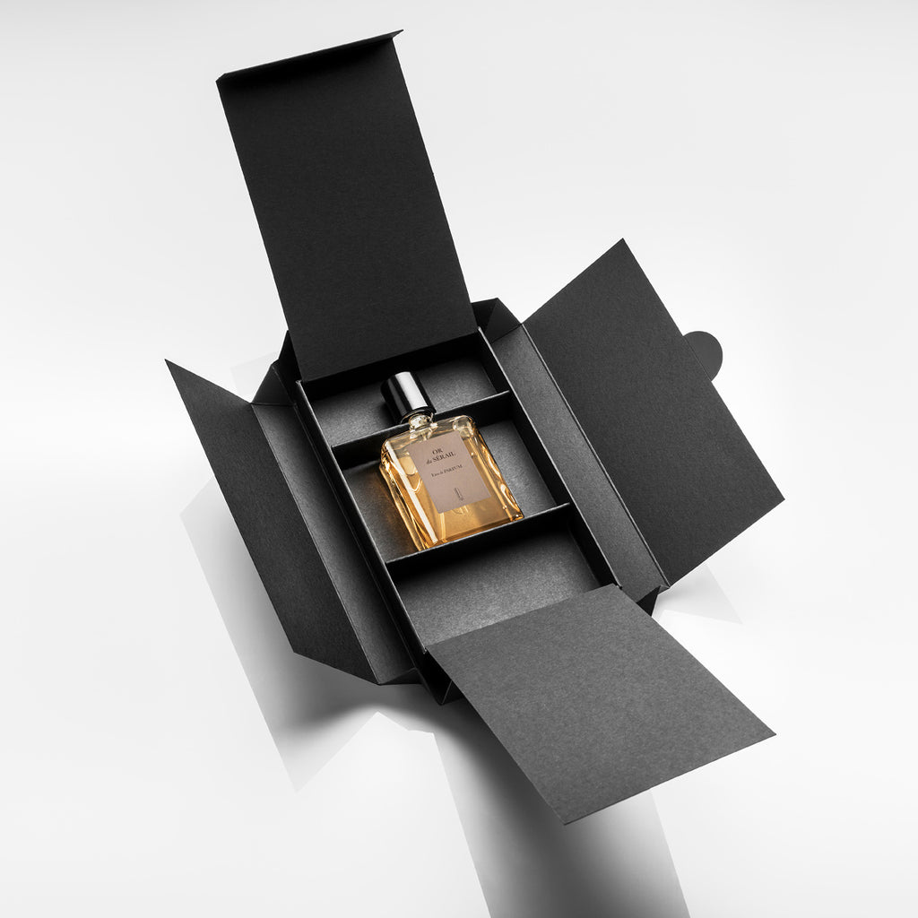 The OR du SÉRAIL perfume by Naomi Goodsir, featuring an amber-colored liquid crafted by Bertrand Duchaufour, is displayed inside an opened black cardboard box on a white background.